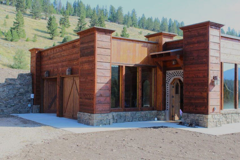 Montana Earthship entry facade clad in wood with a bottle-wall arch door, hills behind