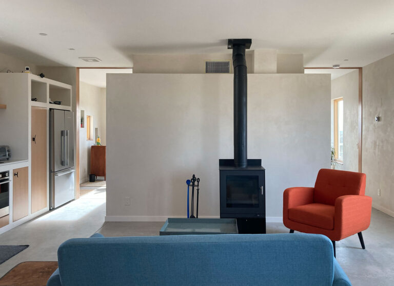 Living room with a wood stove, plaster walls and kitchen beyond